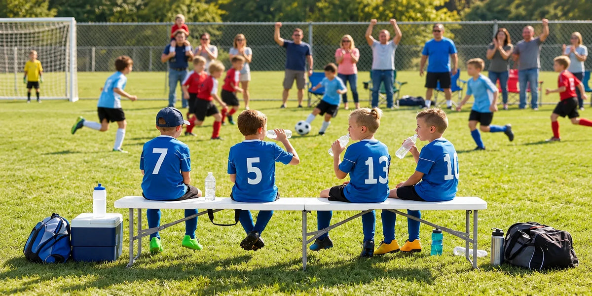 Four young soccer players sitting on a white 6FT folding bench during a soccer match, with water bottles and sports bags beside them.